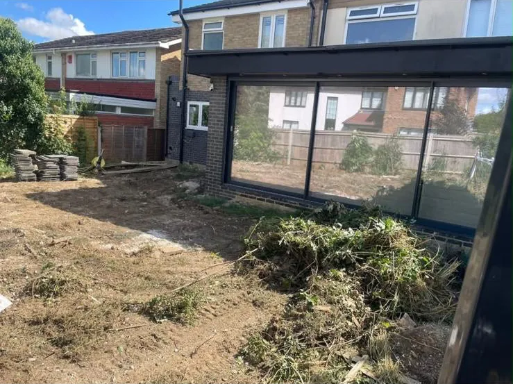 Before: a rear garden stripped back to bare earth, cut greenery piled up and paving slabs stacked ready, beside a dark grey extension with sliding doors