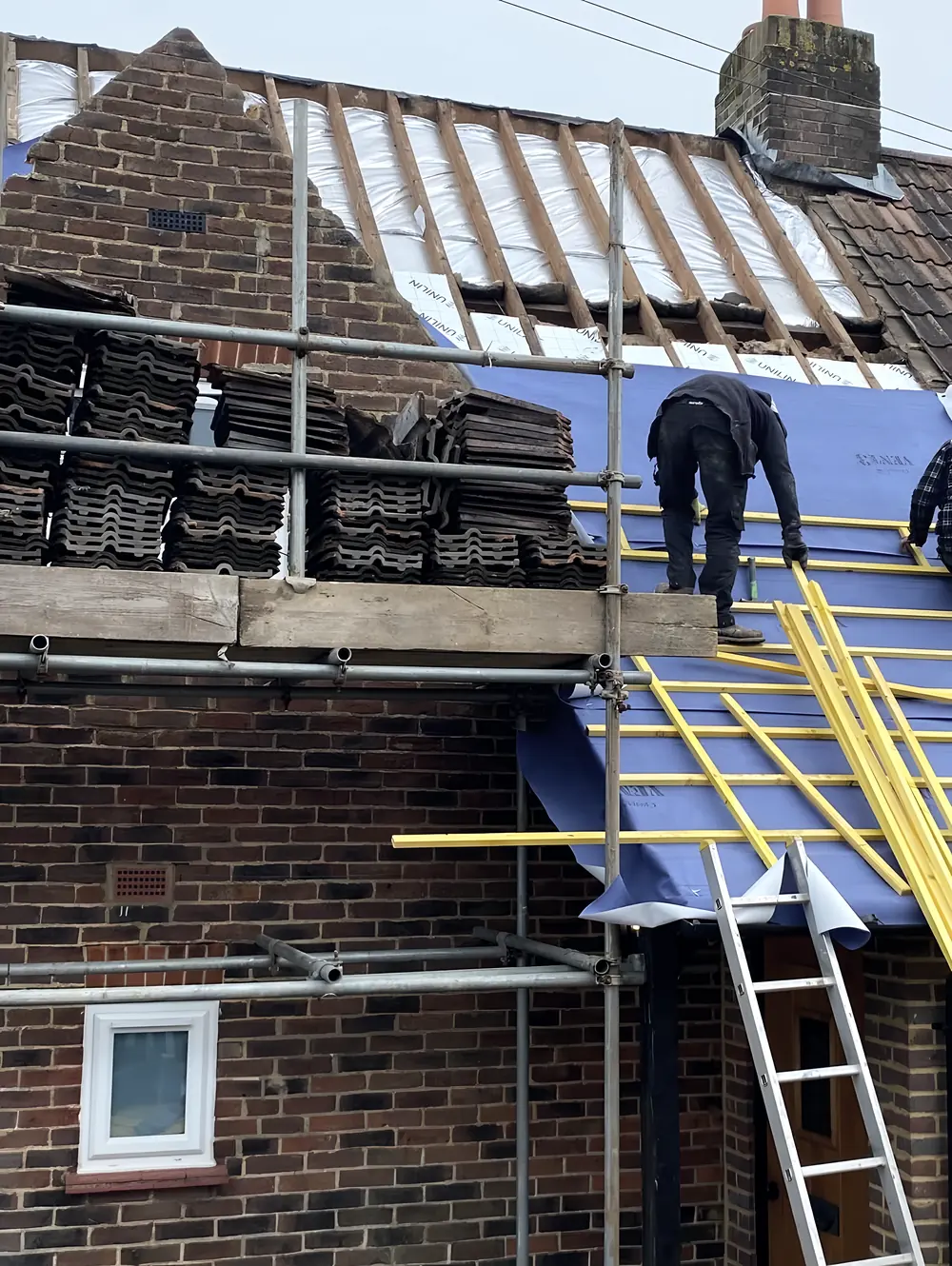 A roofer on blue membrane with new battens, old tiles stacked on the scaffold beside him