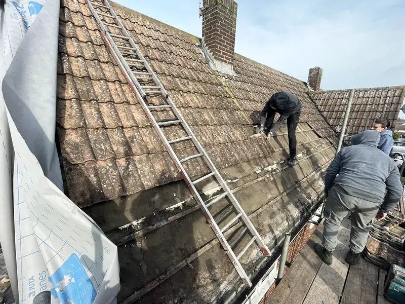 Roofers on a tiled slope, the lower courses stripped back to the battens, a ladder laid up the roof