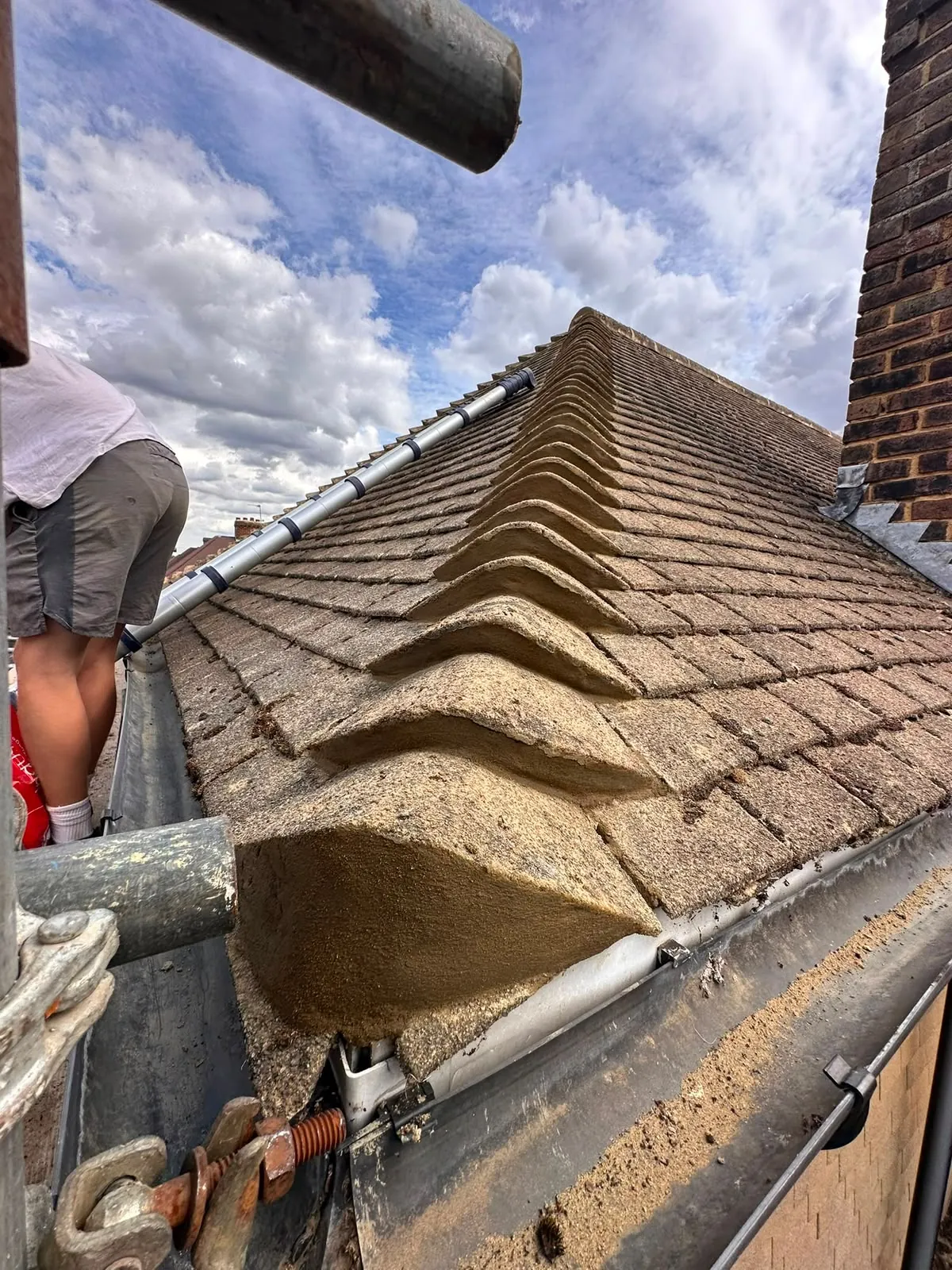 Hip of a tiled roof bedded in fresh mortar, with scaffold alongside and a brick chimney stack to the side