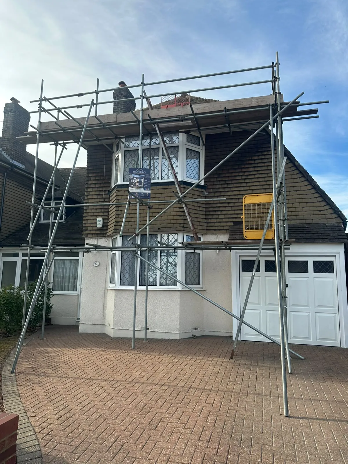 Scaffold erected across the front of a tile hung house with the roof stripped back along the ridge and a roofer working at the top