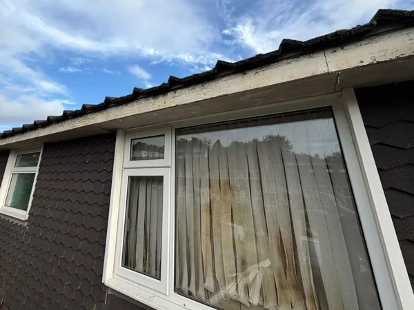 Before: old painted fascia and guttering along the eaves of a house with hexagonal tile cladding, over a big window with vertical blinds