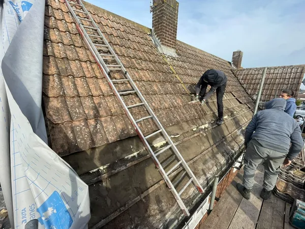 Roofers on a tiled slope, the lower courses stripped back to the battens, a ladder laid up the roof